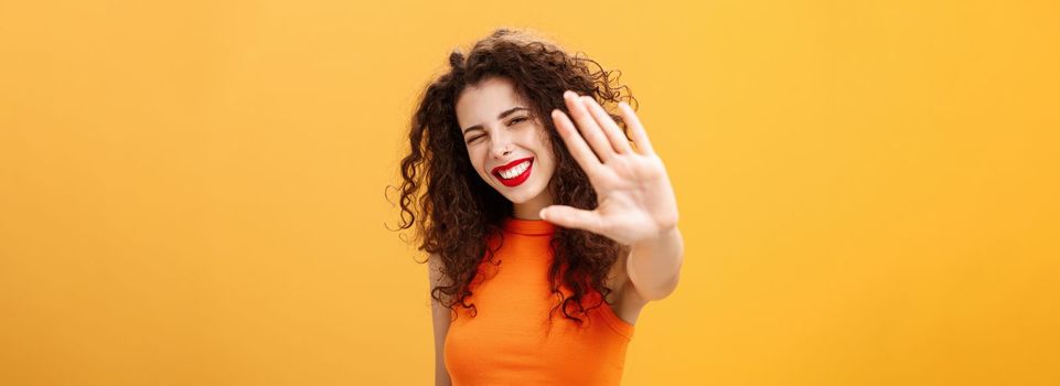 Give me five mate. Stylish friendly and joyful attractive woman with curly hairstyle winking and smiling happily pulling hand towards camera to greet or congratulate friend over orange background. Copy space