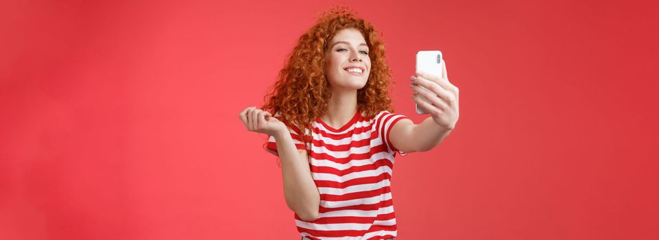 Lifestyle. Cute pleased happy good-looking redhead ginger girl curly hairstyle smiling perfect toothy grin posing delighted amused hold smartphone taking selfie record video message tenderly red background.