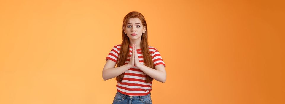 Silly redhead innocent girl acting pity, pulling sad face begging for help, hold hands pray, frowning, purse lips upset, plead favor, apologizing, saying sorry, stand orange background in need.