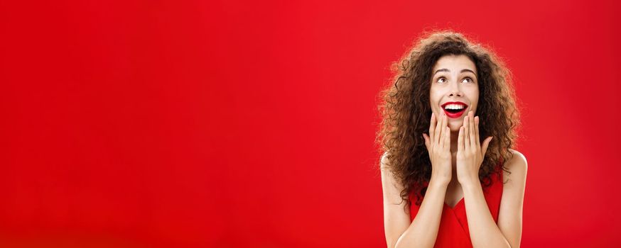 Waist-up shot of amused and entertained happy young dreamy girlfriend. in stylish dress holding palms on chin and opening mouth with broad smile looking delighted up as if seeing miracle over red wall.