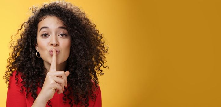 Shh please not tell secret. Cute and tender woman with curly hairstyle gently shushing with index finger on folded lips asking keep promise and remain silent, making surprise over yellow wall.