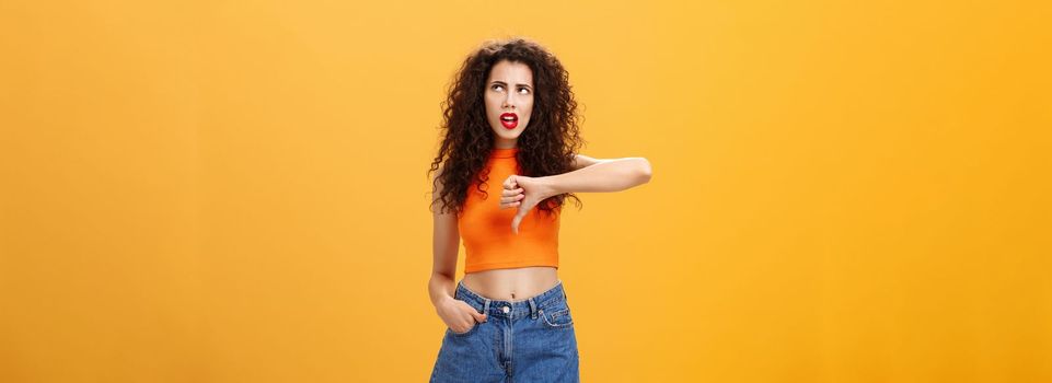 Party for losers I am out. Dissatisfied attractive popular girl with curly hairstyle in red lipstick and cropped top rolling eyes up from irritation and dislike showing thumb down over orange wall.