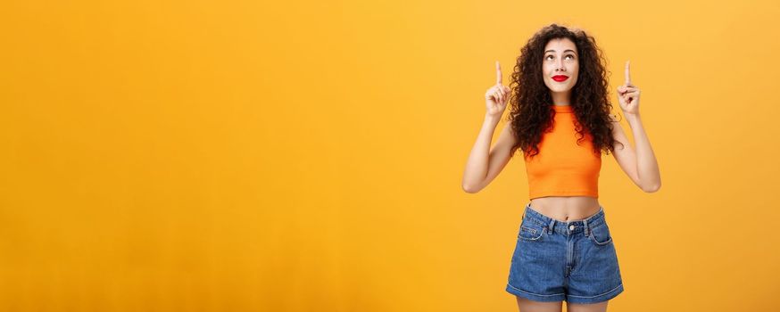 Woman wishing on shooting start believing in miracle looking and pointing hopefully up dreaming and praying in plan fullfillment standing in stylish urban cropped top over orange background. Copy space