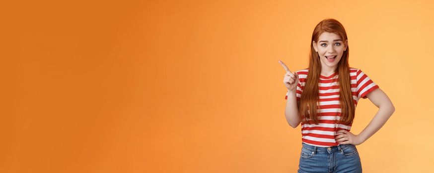 Lively friendly redhead woman discuss new trend pointing upper left corner, smiling, talking joyfully, hold hand chest relaxed casual pose, stand orange background cheerful show advertisement.