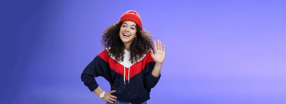 Hey nice to meet you. Charming female ski couch in cute red beanie with curly hair waving hello with palm and smiling broadly greeting newbies as teaching winter sports over blue background. Copy space