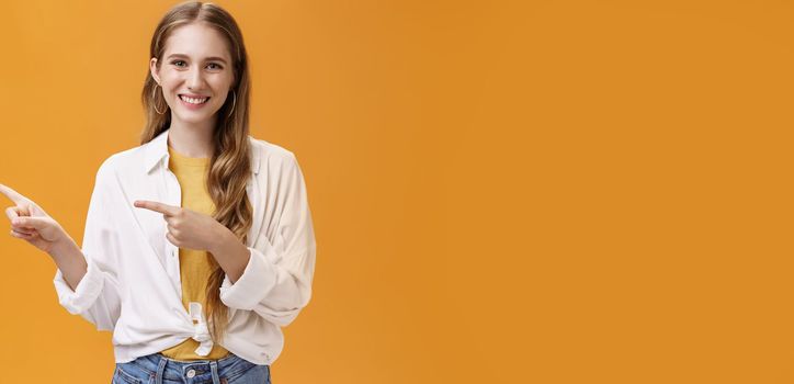 Lifestyle. Waist-up shot of friendly pleasant and stylish young teenage female in blouse over t-shirt with cute wavy fair hair pointing left and smiling broadly at camera posing happy against orange background.
