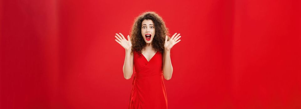 Passionate amazed and thrilled european adult woman in evening dress with curly hairstyle screaming from delight and surprise waving with raised hands and opening mouth from amazement over red wall. Copy space