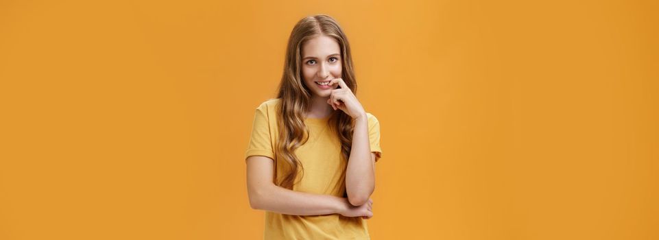 Creative tricky young woman with natural wavy long hair in yellow t-shirt looking from under forehead with intention and lust in expression biting finger, smiling at camera over orange wall.