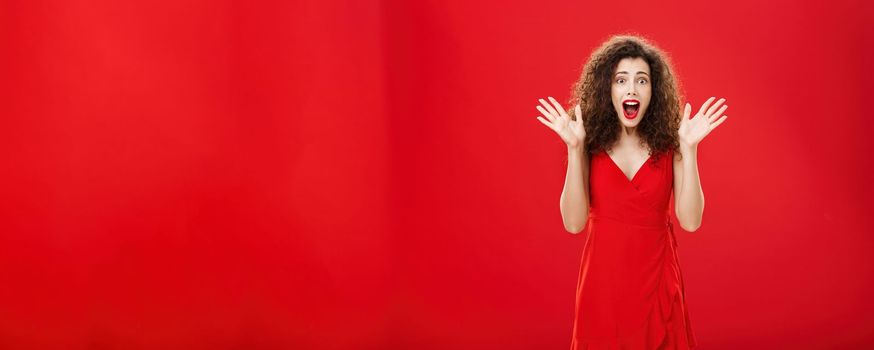 Passionate amazed and thrilled european adult woman in evening dress with curly hairstyle screaming from delight and surprise waving with raised hands and opening mouth from amazement over red wall. Copy space