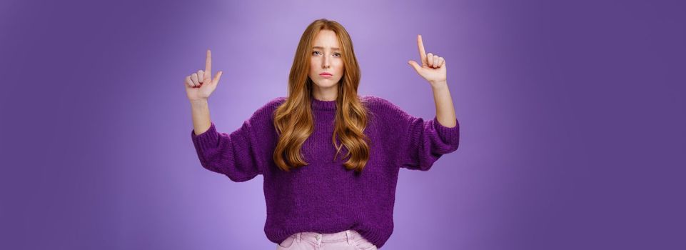 Disappointed gloomy and moody attractive redhead woman with freckles in warm sweater raising hands pointing up and reacting with sadness and regret on product or copy space, being unsatisfied, unhappy.