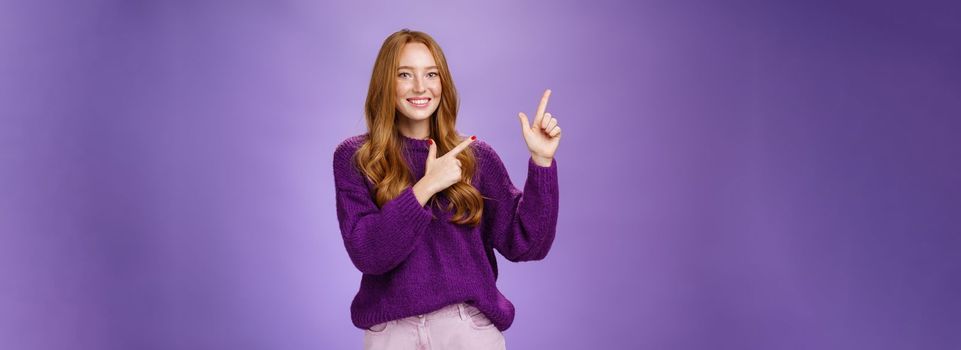 Lifestyle. Charming assertive female shop assistant pointing at upper left corner to promote cool product smiling broadly feeling joyful and excited expressing friendly attitude as posing in purple sweater.