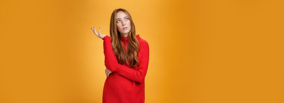 Portrait of unsure thinking redhead girlfriend in red sweater looking uncertain at upper left corner, recalling, feeling nostalgic, solving puzzle in mind, posing against orange background. Copy space
