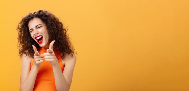Hey what is up. Friendly and outgoing attractive curly-haired female with red lipstick pointing at camera with finger guns as if greeting or hinting friend winking at camera over orange background.