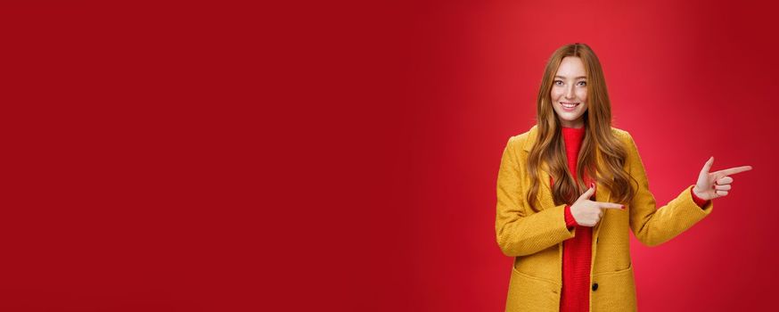 Carefree cute and tender young redhead female student in stylish yellow coat over dress pointing right and smiling with delighted and impressed look showing great place over red background. Advertisement, promotion concept