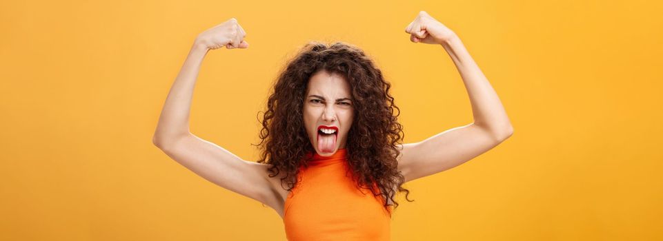 Waist-up shot of cool and daring caucasian female in orange top with tattoo on arm frowning making funny face sticking out tongue raising hands showing muscles feeling power and strengths. Copy space