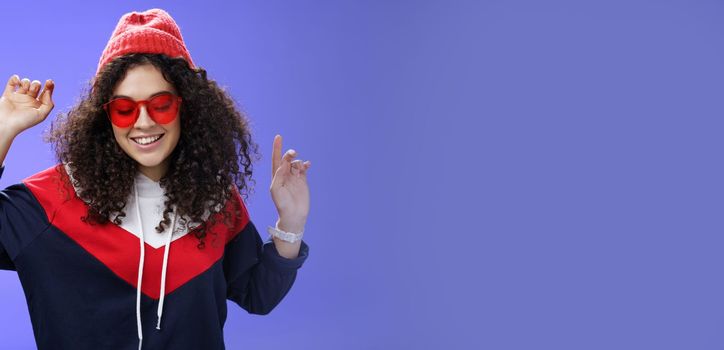 Girl feeling awesome having fun and enoying cool party, dancing with raised hands looking down smiling broadly wearing sunglasses and stylish beanie posing against blue background.