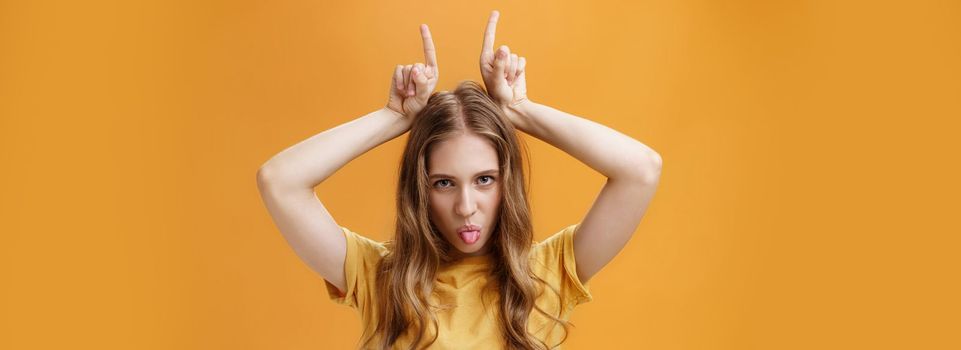 Daring and cool emotive attractive female student with wavy fair hair and little scars showing tongue rebellious looking from under forehead holding index fingers on head being stubborn. Copy space