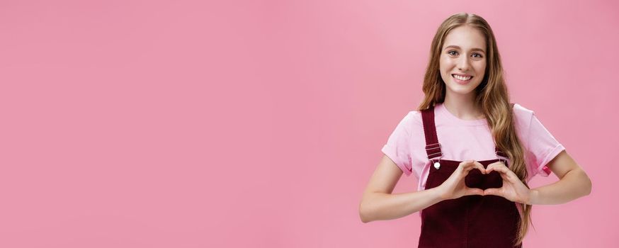 Girl loves family. Kind charming young woman in overalls with small tattoo on arm showing heart gesture over body and smiling lovely at camera expressing tender and cute attitude over pink wall.