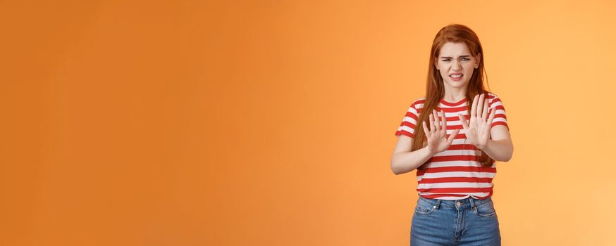 Gosh it stinks. Disgusted redhead picky woman blocking sign raise hands up defensive, grimacing, cringe from aversion awful smell, show refusal rejecting disgusting offer, stand orange background.