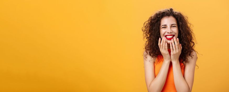 Waist-up shot of delighted charming and cute curly-haired caucasian female. in cropped top being touched with sweet words smiling pleased holding palms on jaw-line surprised and happy over orange wall.