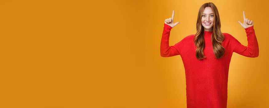 Enthusiastic and excited energized redhead female in red warm sweater pointing up with rised hands and smiling broadly thrilled with awesome promotion, feeling astonished against orange background.
