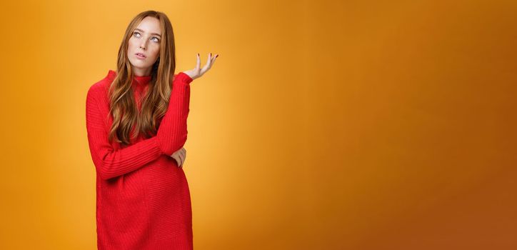 Portrait of unsure thinking redhead girlfriend in red sweater looking uncertain at upper left corner, recalling, feeling nostalgic, solving puzzle in mind, posing against orange background. Copy space