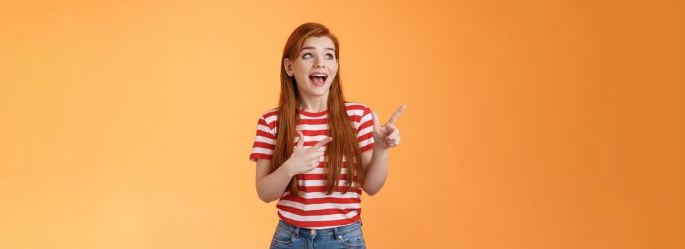 Enthusiastic cheerful redhead woman lively smiling laughing joyfully, enjoy watching copy space, look pointing upper left corner joyful, excited see awesome advertisement, orange background.