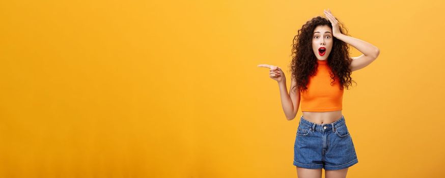 Surprised thrilled beautiful caucasian girl. with curly hairstyle in red lipstic and orange cropped top holding hand on head amazed and pointing left questioned and astonished over studio background.