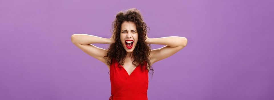 Ex-boyfriend spoilt woman formal event being outraged. and pissed yelling in fury holding hands on curly hair grimacing from anger standing over purple background in stylish evening red dress.