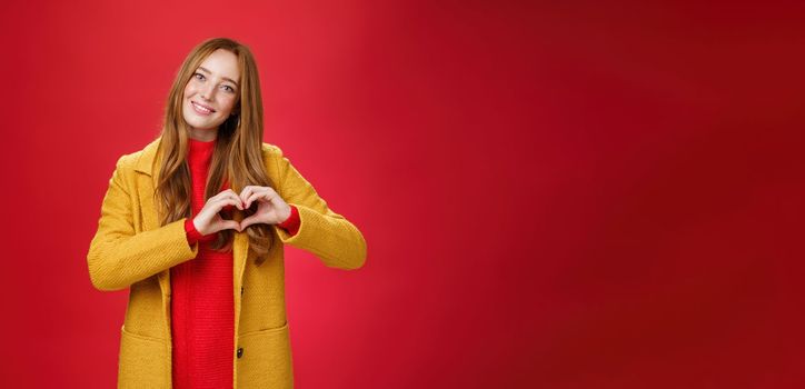 Love, romance and fall concept. Portrait of charming tender and gentle young redhead woman in yellow coat showing heart gesture making confession in sympathy, smiling cute at camera over red wall.