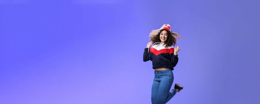 Lifestyle. Delighted carefree and happy young woman with curly hair in warm beanie jumping joyfully having fun raising hands and smiling broadly posing over blue background in outdoor clothes.