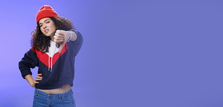 Nah dislike. Portrait of annoyed and unimpressed arrogant cool girl with curly hair tilting head with distain and scorn showing thumbs down in disappointment grimacing, posing over blue wall. Copy space