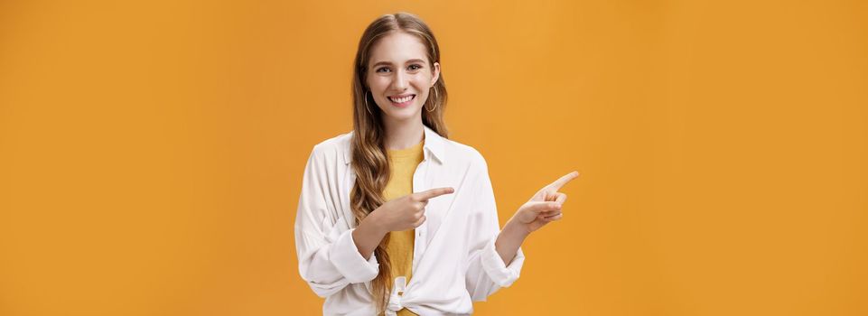 Lifestyle. Waist-up shot of friendly pleasant and stylish young teenage female in blouse over t-shirt with cute wavy fair hair pointing left and smiling broadly at camera posing happy against orange background.