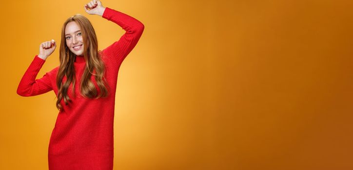 Joyful and carefree relaxed ginger girl in red warm dress partying having fun enjoying cool music as dancing with raised hands smiling broadly at camera against orange background. Lifestyle, people, winter concept