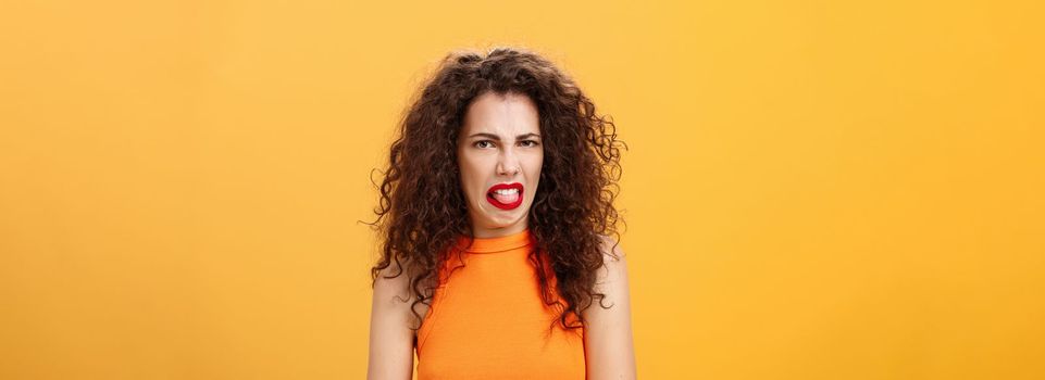 Girl being disgusted with row food sticking out tongue and frowning showing aversion and disgust talking about thing she dislikes with friend standing intense and dissatisfied over orange background. Copy space