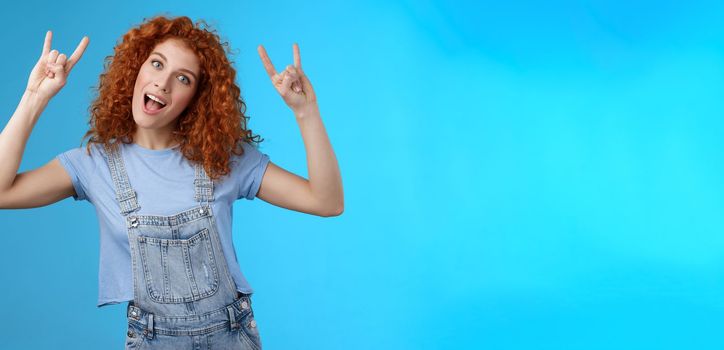 Lifestyle. Daring cool stylish awesome redhead cheerful curly-haired girl tilt head show tongue joyfully stare camera playful raise hands rock-n-roll heavy metal gesture having fun blue background.