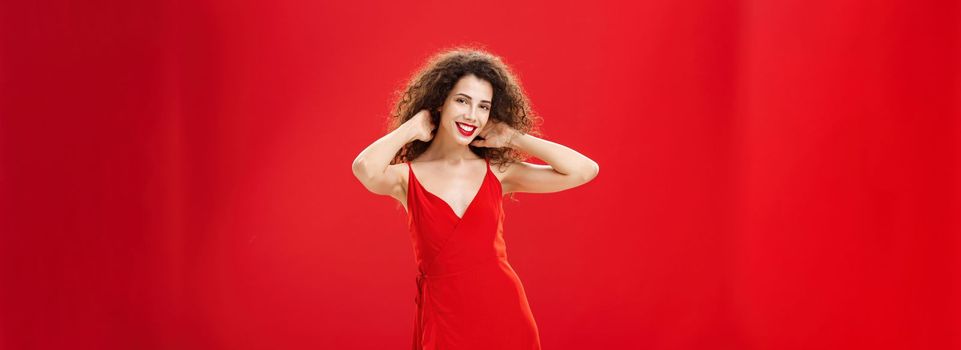 Stylish gentle caucasian girl with curly hairstyle and evening make-up touching strands of hair checking haircut and smiling broadly at camera posing in new elegant red dress over studio background. Copy space