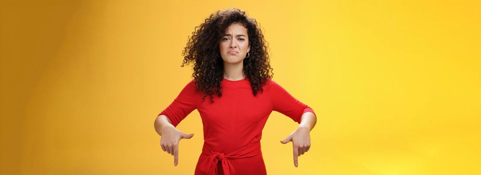 Gloomy disappointed cute silly curly-haired female in red dress frowning upset and making sad smile as pointing down with regret and unsatisfied expression, jealous or displeased over yellow wall.