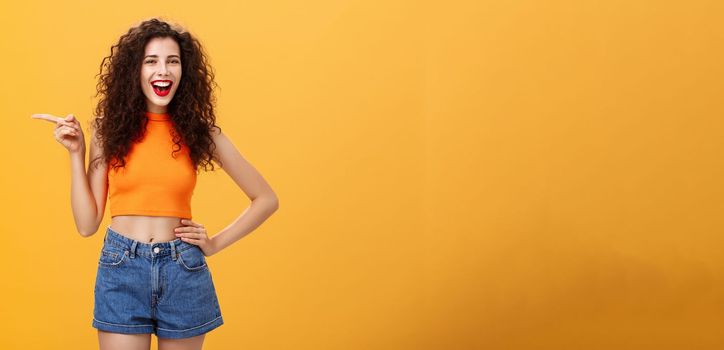 Girl suggesting check out cool copy space. Attractive young happy and energizing curly-haired female in cropped top and denim shorts holding hand on waist pointing left and laughing joyfully. Directions and advertisement concept