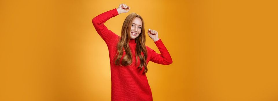 Joyful and carefree relaxed ginger girl in red warm dress partying having fun enjoying cool music as dancing with raised hands smiling broadly at camera against orange background. Lifestyle, people, winter concept