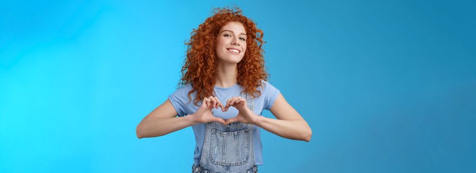 Sassy confident good-looking modern redhead curly woman raise head proud love tell girlfriend romantic heartwarming feelings present own heart smiling broadly standing blue background.