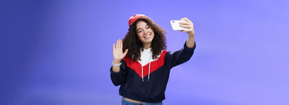 Studio shto of stylish cute female vlogger with curly hair in winter red hat smiling and waving hello at smartphone camera as recording video, making interesting content to post online over blue wall.