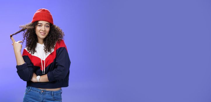 Stylish and snobbish arrogant curly-haired woman in warm beanie rolling curl on finger smirking and looking with contempt at camera, scorning person as being too cool over blue background.