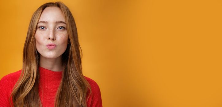 Close-up shot of flirty good-looking sensual redhead female in red knitted clothes folding lips in kiss and gazing at camera as giving mwah feleing romantic and pretty against orange background. Body language, fashion and people concept