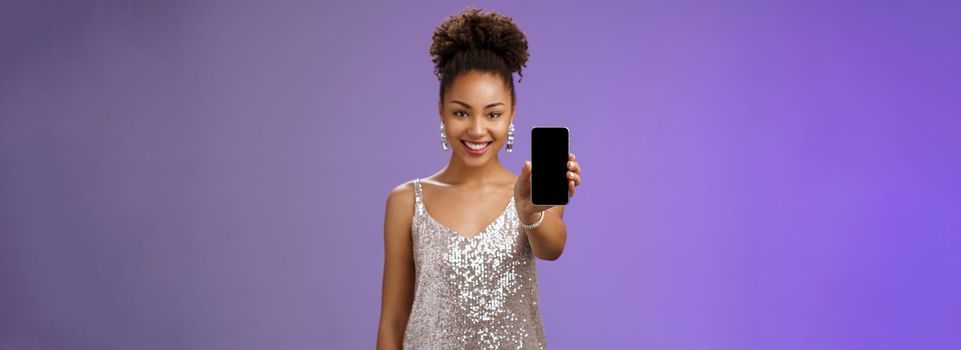 Charming attractive african-american woman in silver glittering stylish dress extend arm showing smartphone display presenting app promoting cool device standing pleased smiling blue background.