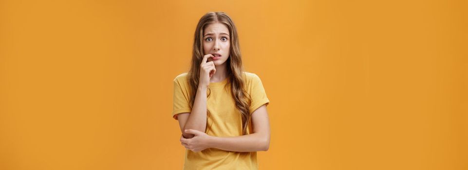 Girl worried mom react on new tattoo. Portrait of anxious and nervous silly insecure young female with wavy natural hairstyle biting finger panicking looking concerned and troubled over orange wall. Emotions concept