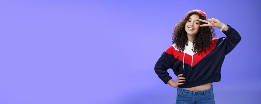 Lifestyle. Attractive feminine woman with curly hairstyle in hat and sweatshirt showing peace or victory sign around eye and smiling carefree having fun playing in yard with snow over blue background.