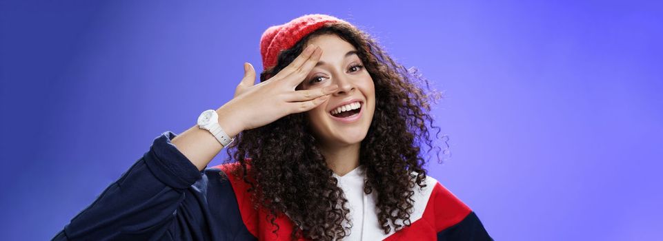 Close-up shot of charismatic happy carefree woman in winter red cute beanie and sweatshirt holding fingers near eye and peeking with broad smile at camera having fun, playing over blue background.