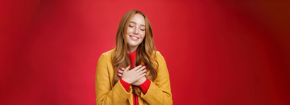 Girl feeling like heart warming. as remembering sweet tender memories with closed eyes and silly broad smile holding hands on chest pressing to soul posing nostalgic and touched over red background.