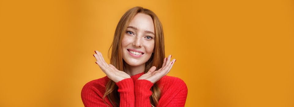 Close-up shot of sensual tender and gentle ginger girl. with romance and happy gaze holding palms near pure clean skin with freckles smiling joyfully tilting head looking friendly over orange wall.